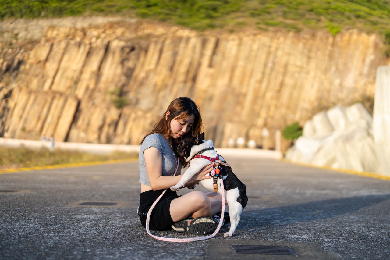 A young woman sits with her French bulldog on a sunny day against a rocky backdrop.